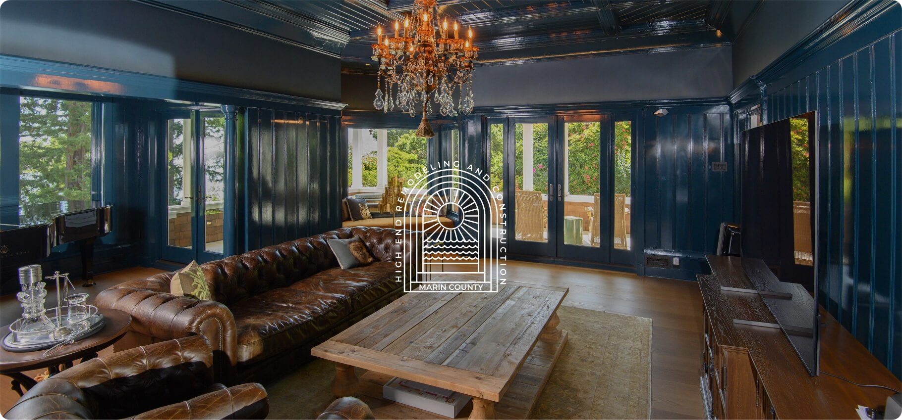 A luxurious living room with a dark blue theme, featuring a chandelier, a leather sofa, and large windows.